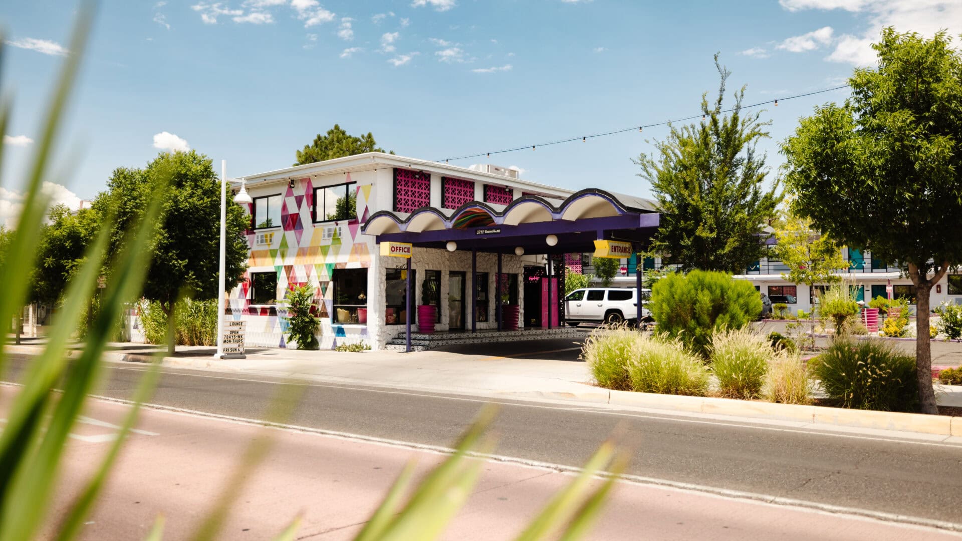 Two-story roadside hotel with maroon trim and a covered entrance along Route 66 in Albuquerque, New Mexico, under a blue sky with tall grass in the foreground.